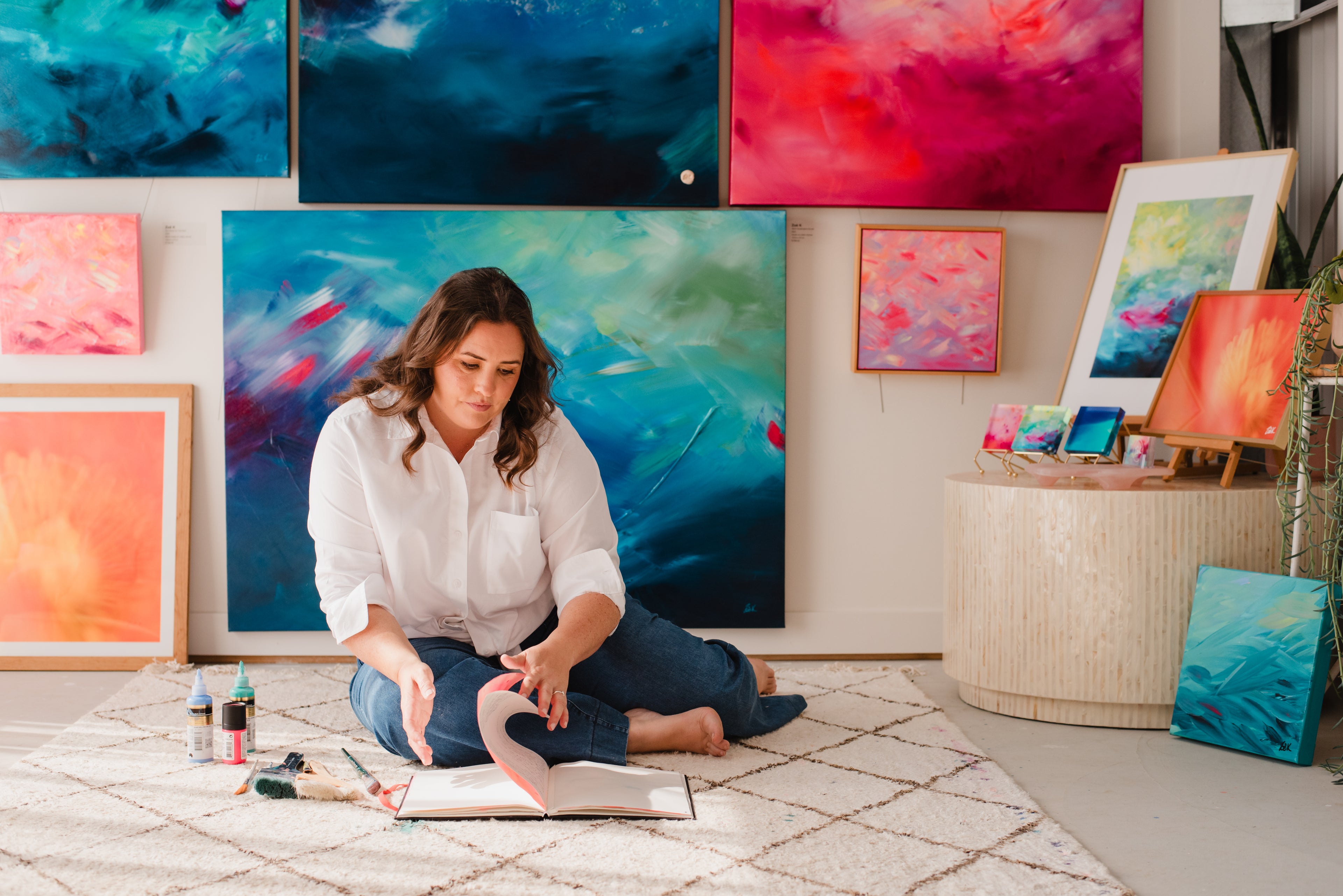 Woman sitting on the floor surrounded by colorful abstract paintings in a gallery setting.