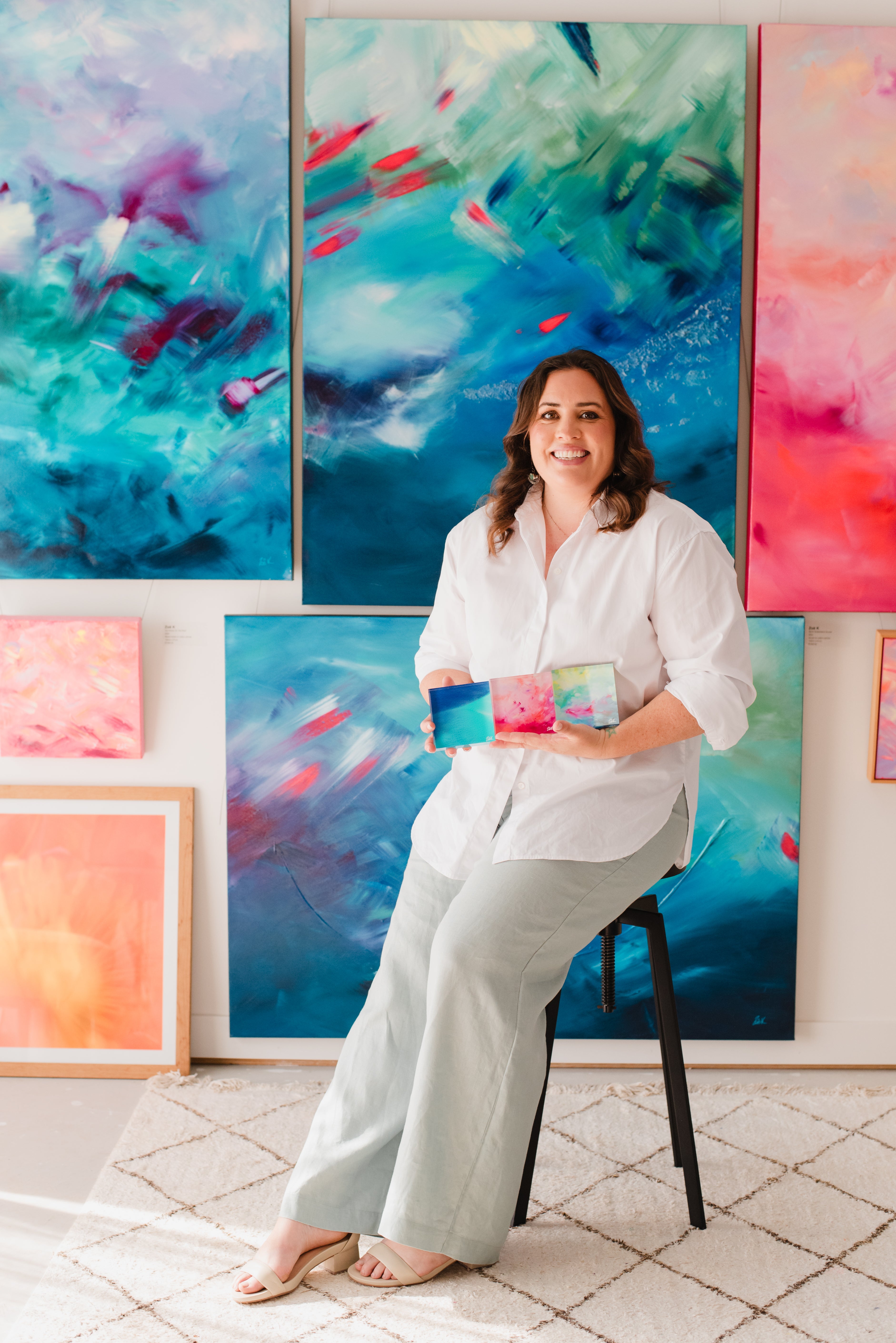 Art for your home. Woman holding a small painting in front of colorful abstract artworks